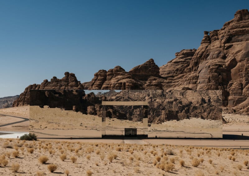 A large, rectangular building with mirrored walls reflects the surrounding desert landscape and rocky cliffs under a clear blue sky. Sparse shrubs dot the sandy ground in front of the structure.