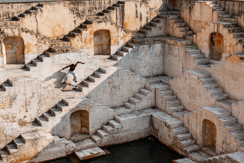 A skateboarder performs a trick on the steps of an ancient, weathered stepwell with geometric patterns and multiple staircases, set against a backdrop of stone walls and water at the bottom.