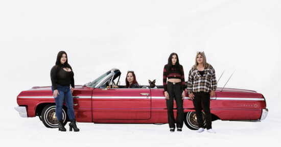 Four women stand next to a red vintage convertible car with gold rims, while a dog sits in the driver's seat. The background is plain white.
