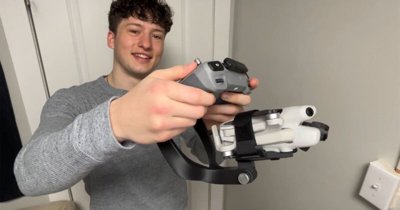 Un joven de pelo rizado y camisa gris sostiene un dron y su controlador y sonríe a la cámara en una habitación con paredes claras y una puerta blanca al fondo.