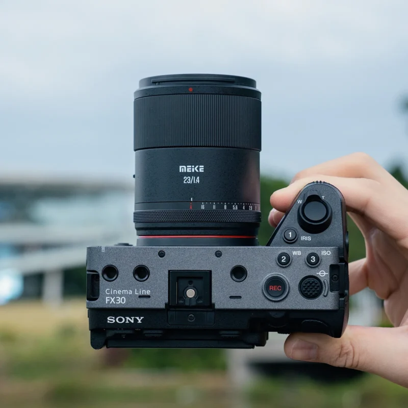 A hand holds a Sony Cinema Line FX30 camera with a Meike 23mm f/1.4 lens, showing the top view of the camera against a blurred outdoor background.