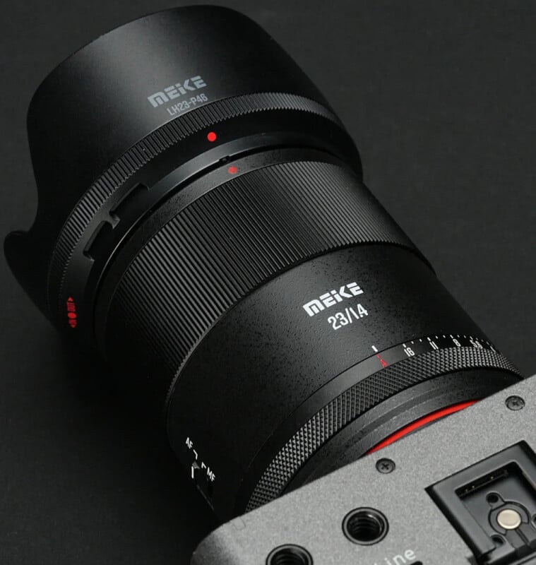 A close-up of a Meike 23mm f/1.4 camera lens attached to a camera body, placed on a black background. The lens details and branding are clearly visible.