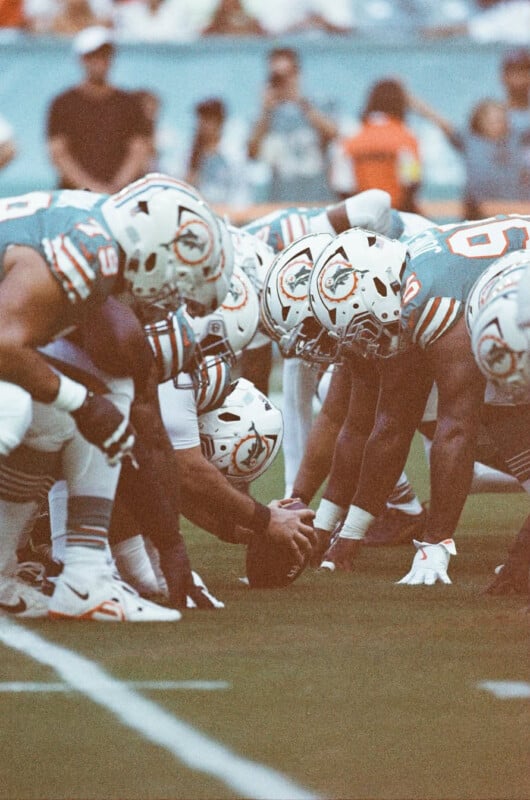 Two American football teams face off at the line of scrimmage, ready for the snap. Players in teal jerseys and white helmets with a dolphin logo prepare for the play on a grassy field.