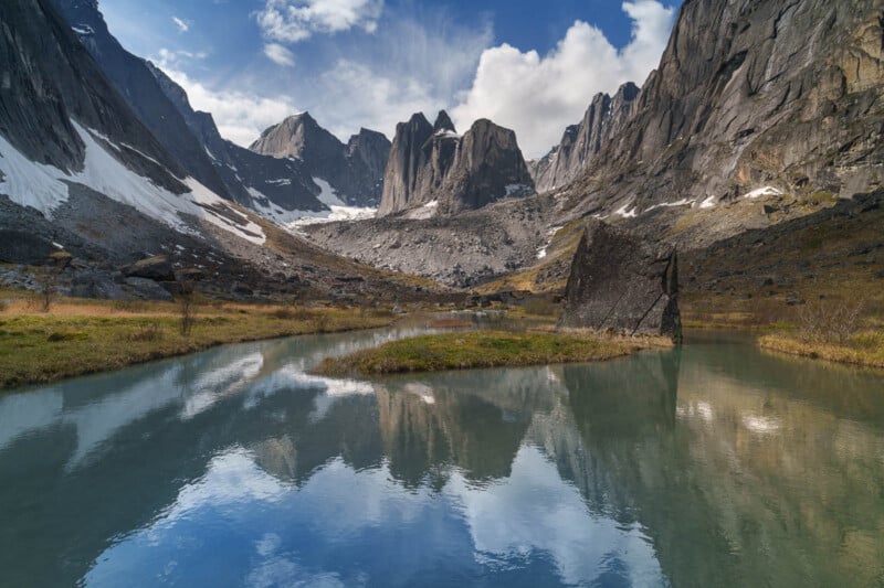 A clear stream reflects jagged mountain peaks, rocky slopes, and partly cloudy sky with patches of snow and grass in a dramatic alpine valley landscape.