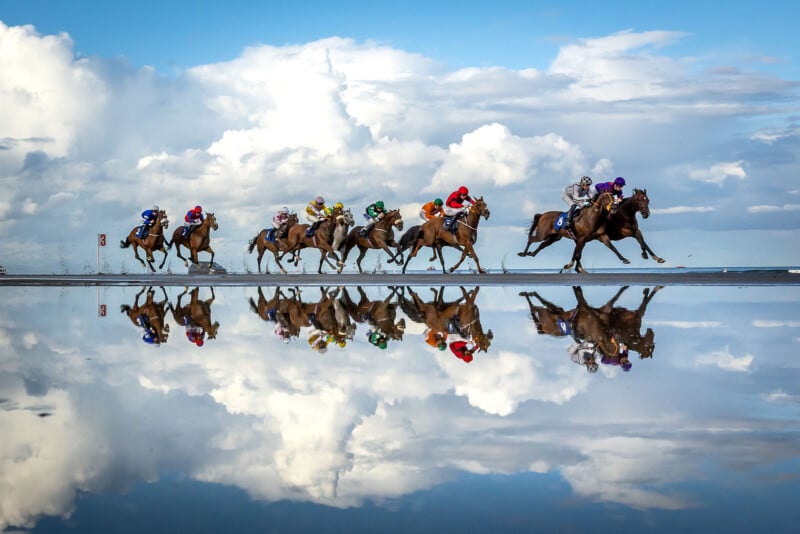 A group of jockeys riding horses race across a wet track, with their reflections clearly visible in the water beneath them against a backdrop of clouds and blue sky.