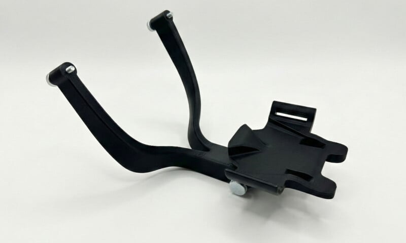 Un soporte de plástico negro en ángulo con dos brazos curvos y una base plana con pequeños sujetadores de metal en el extremo, sobre un fondo blanco sólido.