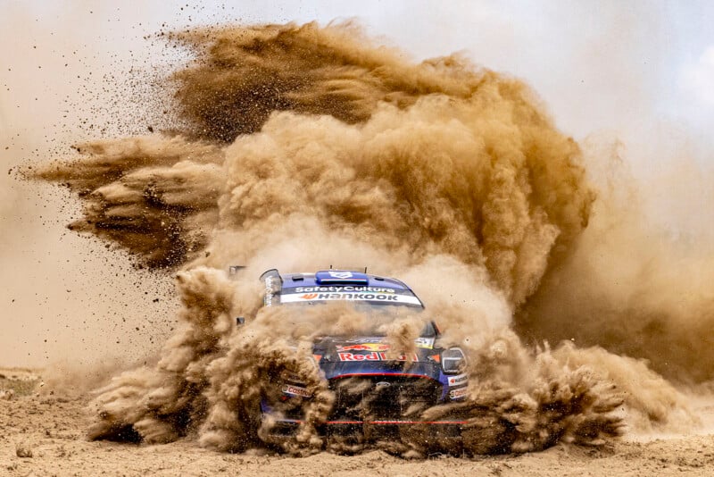 A rally car races through a cloud of dirt and dust, partially obscuring the vehicle as it speeds across a rough off-road track, with debris flying dramatically in all directions.