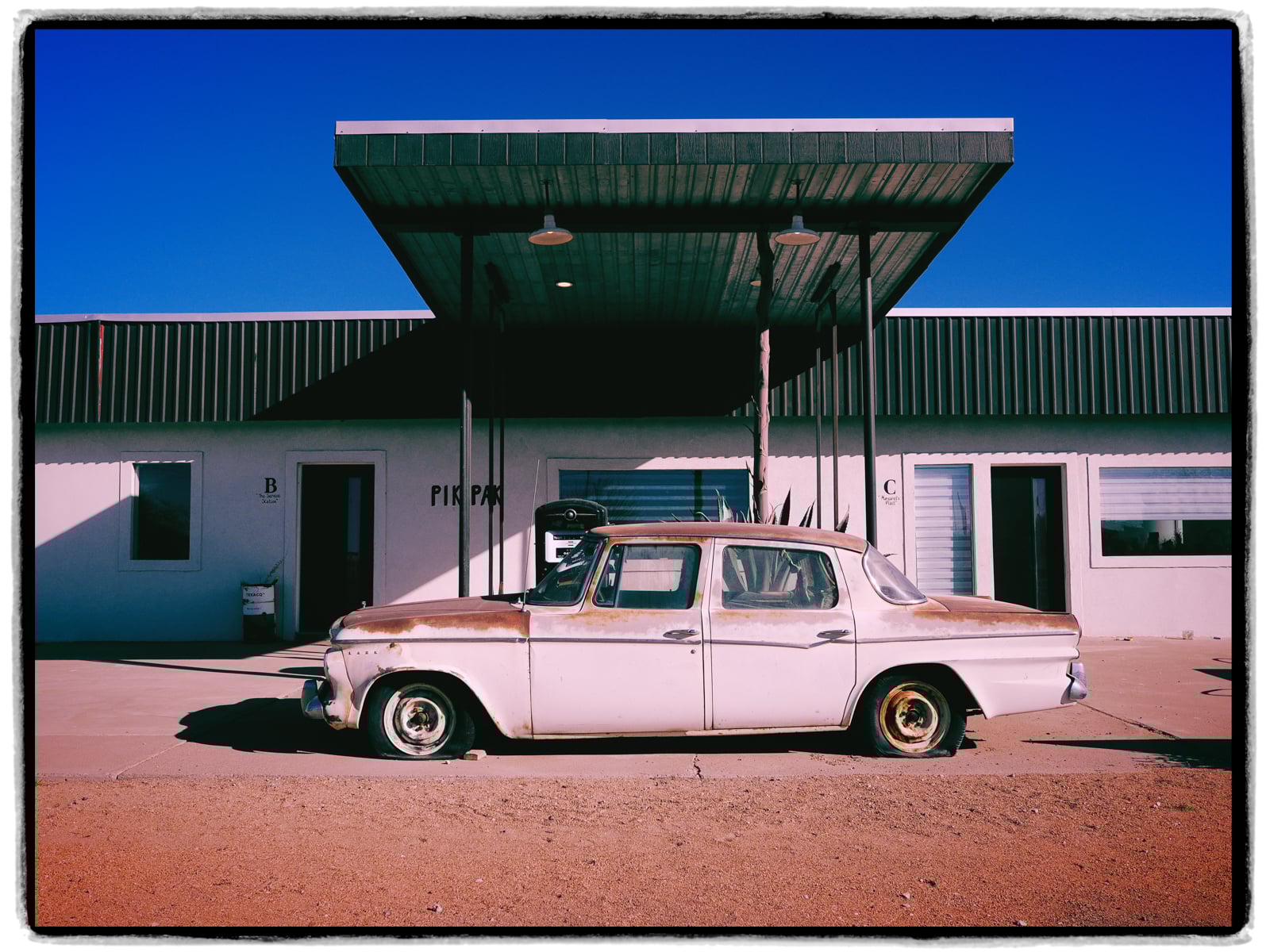 A rusty, vintage white car with flat tires is parked in front of a one-story white building under a metal canopy on a sunny day. The sky is clear and blue.