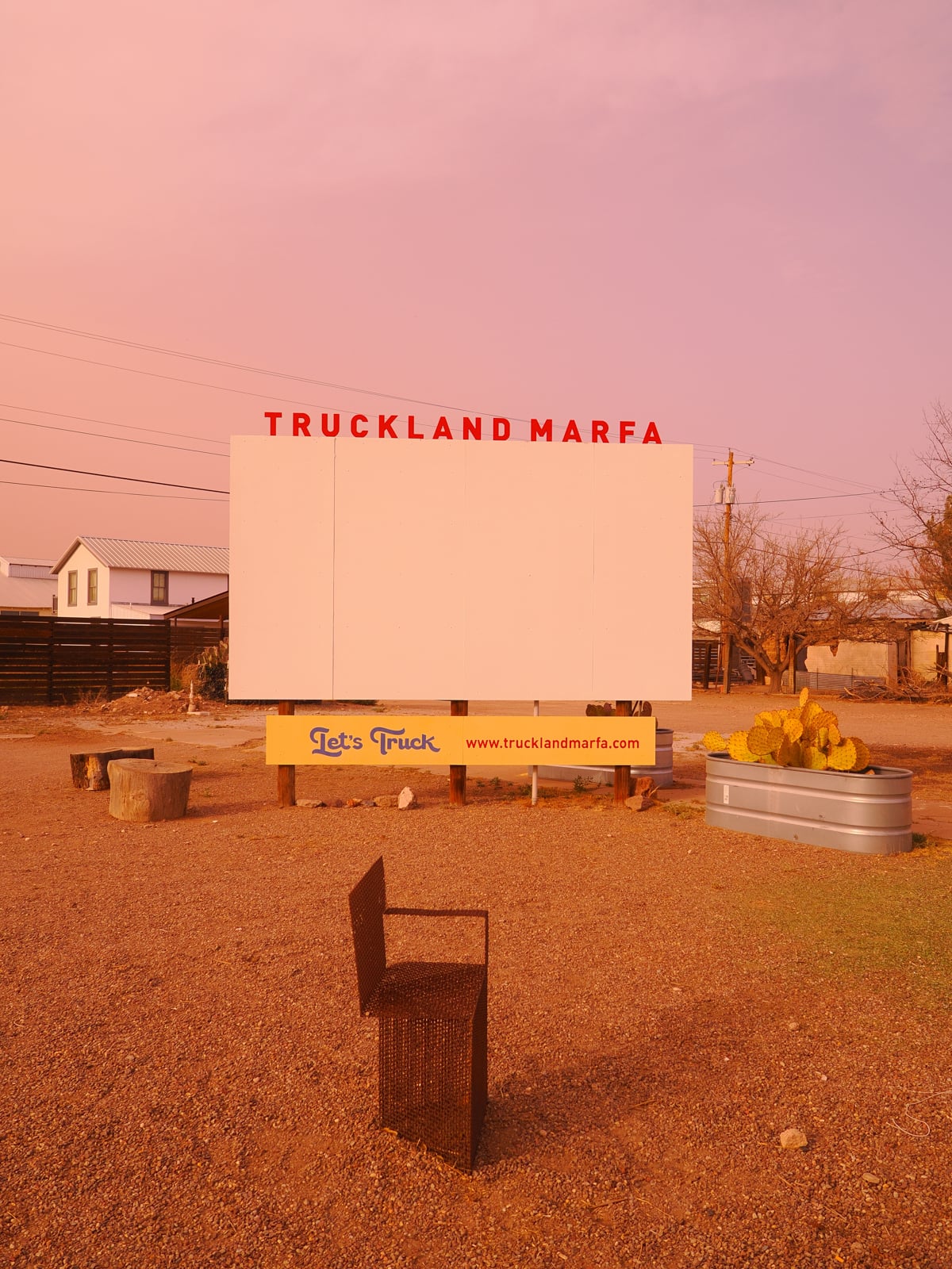A large, blank white billboard with red letters reading "TRUCKLAND MARFA" stands on a dirt lot at sunset. In front, there’s a metal chair, a sign for "Let's Truck," and a metal trough with yellow cacti.
