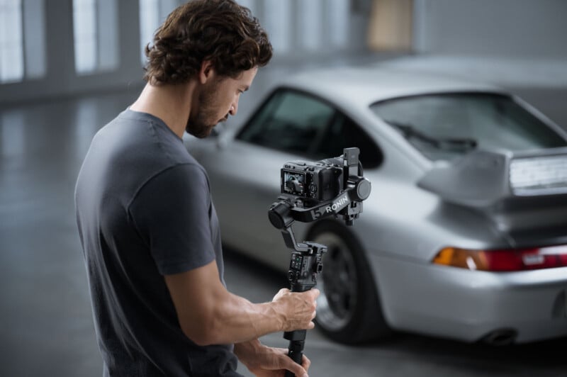 A man holding a camera mounted on a handheld gimbal films a silver sports car in a garage or showroom. The car's rear is visible in the background.