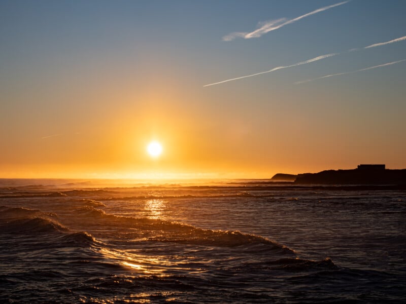 Golden sunset over the ocean with gentle waves, a glowing sun near the horizon, and a silhouetted coastline to the right under a clear sky with faint streaks of clouds.