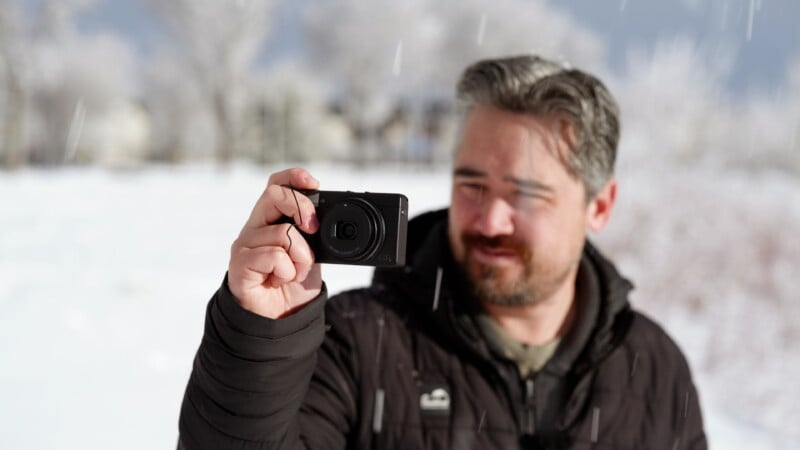 A man with short brown and gray hair, wearing a black jacket, holds a compact camera up and smiles while standing outdoors in a snowy landscape.