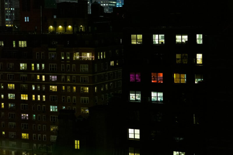 A nighttime view of city apartment buildings with many windows lit up in various colors, including white, yellow, purple, and red, highlighting different rooms and interiors. The surrounding area is dark.