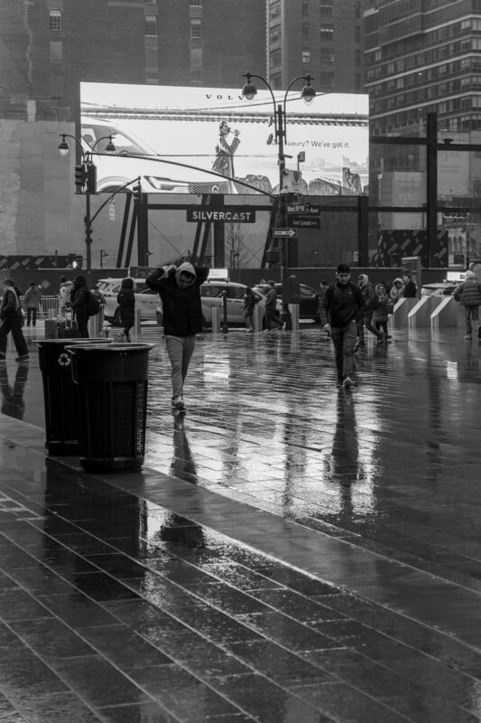 People walk on a wet city street in the rain, some with hoods up. Buildings and a large billboard advertising Volvo can be seen in the background. Reflections appear on the shiny pavement. The image is in black and white.