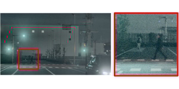 A nighttime street scene with a car approaching a crosswalk where two pedestrians are walking. The pedestrians are highlighted with a red square and shown in a zoomed-in, grainy inset on the right.
