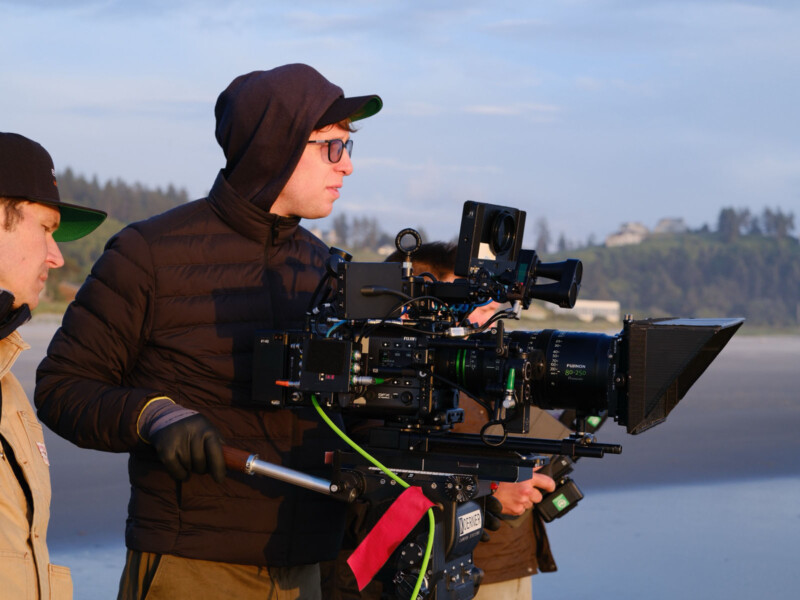 Two people stand on a beach operating a professional film camera. One wears glasses, a hooded jacket, and gloves, focused on filming. Houses and trees are visible in the distant background.
