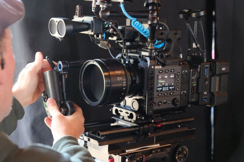 A person operates a professional cinema camera with various attachments and a 100mm lens, adjusting controls while the camera is mounted on a tripod against a dark background.