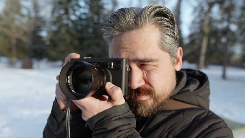 A person with gray hair and a beard is outdoors in a snowy area, holding a camera up to their face and looking through the viewfinder, wearing a dark winter jacket.