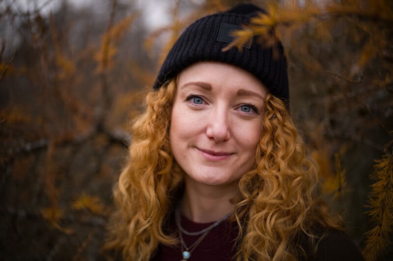 A woman with long curly red hair, wearing a black knit beanie and layered necklaces, smiles softly at the camera. She stands outdoors among branches with yellowish autumn leaves.