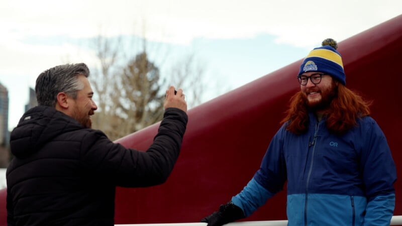 A person in a dark jacket takes a photo of a smiling person with long red hair wearing glasses, a blue winter coat, and a colorful beanie, standing outdoors near a red structure.