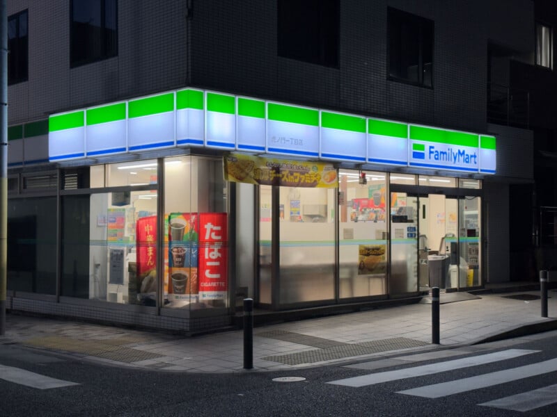A brightly lit FamilyMart convenience store at night, with large glass windows displaying products and colorful advertisements inside. The store sits on a quiet street corner with a sidewalk and bollards in front.