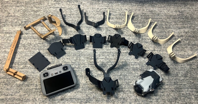 Una variedad de soportes, clips y soportes de plástico e impresos en 3D se alinean sobre una superficie con textura gris, junto a un controlador de drones y un pequeño drone en la esquina inferior derecha.