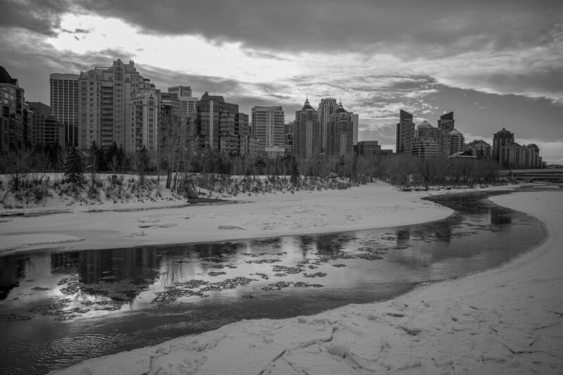 A black and white photo of a city skyline with tall buildings in the background, a partially frozen river in the foreground, and snow covering the riverbanks and trees. The sky is cloudy with sunlight peeking through.