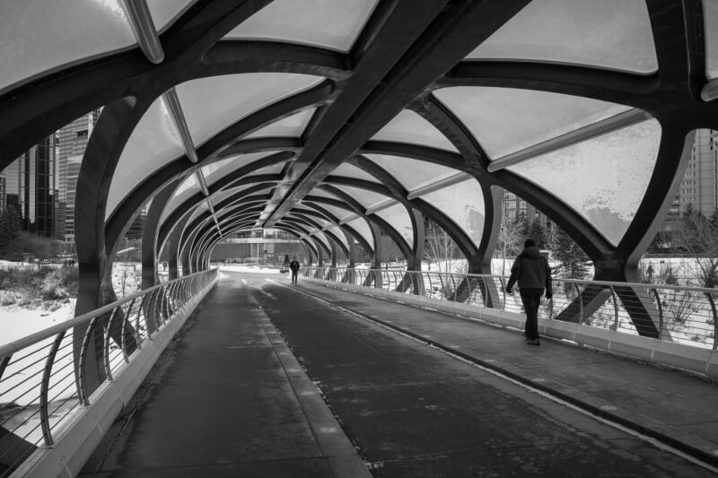 A black and white photo of a modern, covered pedestrian bridge with an arched, geometric design. Two people walk along the pathway, with a city skyline visible in the background on a snowy day.