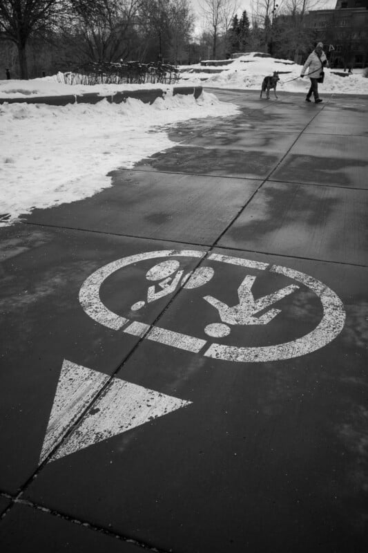 A sidewalk in winter with snow on the ground and a painted pedestrian symbol with an arrow. In the background, a person walks a dog on the path.