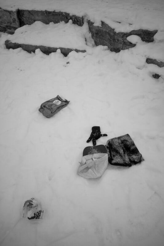 Scattered items, including bags, a glove, and litter, lie on snow-covered ground near a rocky ledge. The scene appears cold and desolate.