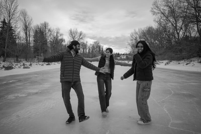 Three people in winter jackets laugh and hold hands while standing on a frozen, snow-covered lake surrounded by bare trees under a cloudy sky.