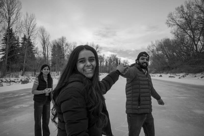 Three people enjoying a winter day on a frozen lake, holding hands and smiling. Bare trees and snow line the background. The image is in black and white.