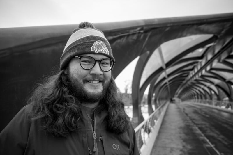 A person with long hair, glasses, and a beanie smiles while standing on a modern, covered pedestrian bridge on a cloudy day. The scene is in black and white.