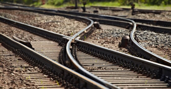 Close-up of railway tracks splitting into multiple directions on a gravel bed, showing a railway switch mechanism under bright daylight.