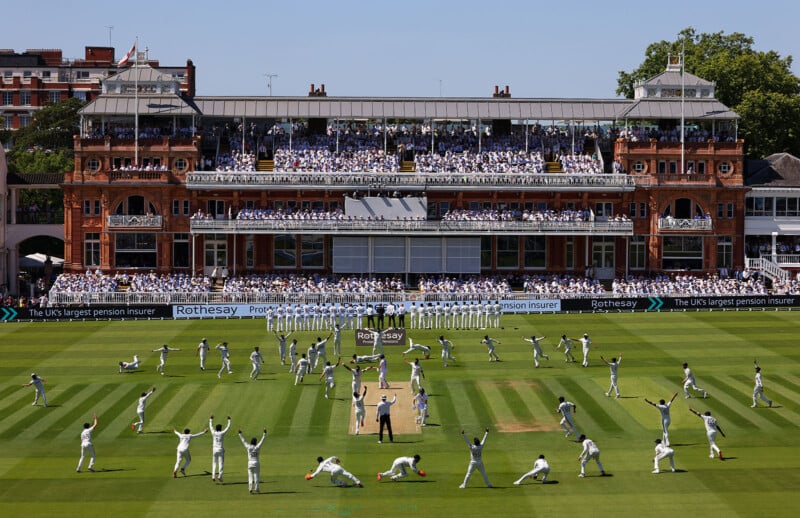 Cricket players on a green field stretching and warming up, with a large historic pavilion and a crowd of spectators in white seated in the background at Lord’s Cricket Ground.