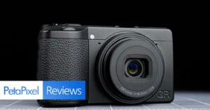 A compact black digital camera is displayed on a grid surface, with "PetaPixel Reviews" text overlaid in the bottom left corner of the image.