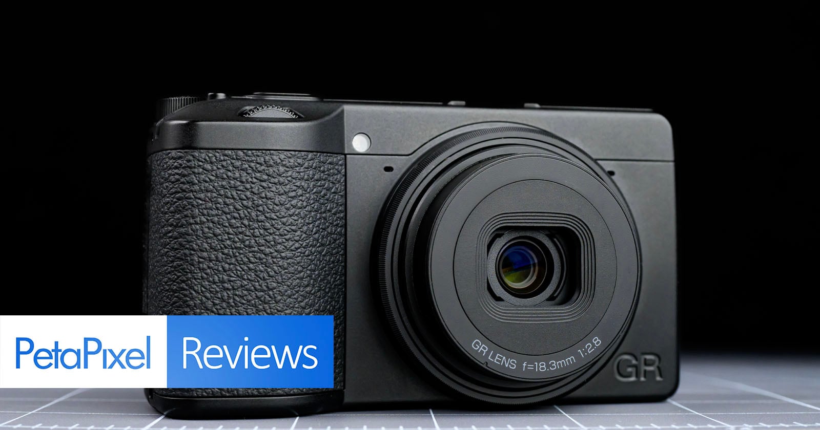 A compact black digital camera is displayed on a grid surface, with "PetaPixel Reviews" text overlaid in the bottom left corner of the image.