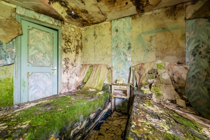 An abandoned, decaying bedroom with mold and moss covering two beds and walls. Wallpaper peels off, paint is chipped, and an old rotary phone sits on a small table between the beds. The ceiling and floor are heavily damaged.