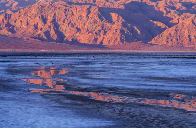 Sunlit mountains glow orange and reflect in a shallow, blue-tinted salt flat. The rocky terrain contrasts with the smooth, rippled surface of the water in the foreground. The scene is serene and bathed in warm, early light.