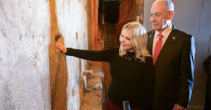 A woman touches an ancient stone wall while a man in a suit stands beside her, both observing the historic site in a dimly lit corridor.