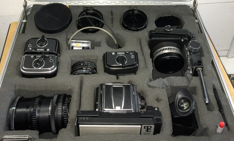 A padded case containing various camera equipment, including camera bodies, lenses, film backs, filters, and accessories, all neatly arranged in custom-cut foam compartments.