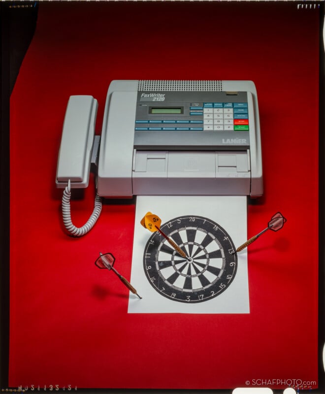 A fax machine and phone rest on a bright red surface. A printed dartboard is placed beneath them, with three darts stuck into the paper, one hitting the bullseye.