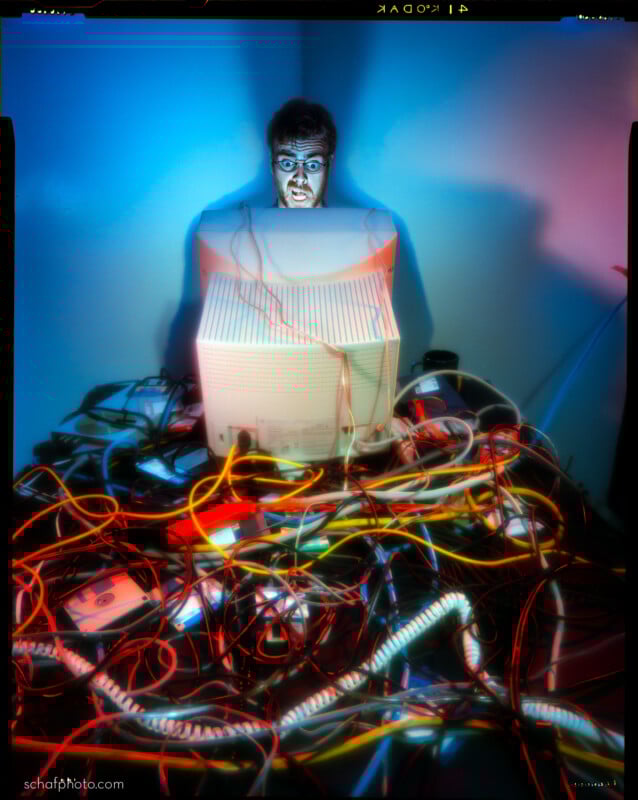 A person with a surprised expression sits behind a vintage computer monitor, surrounded by a chaotic tangle of colorful cables and electronic devices, lit by dramatic blue and red lighting.