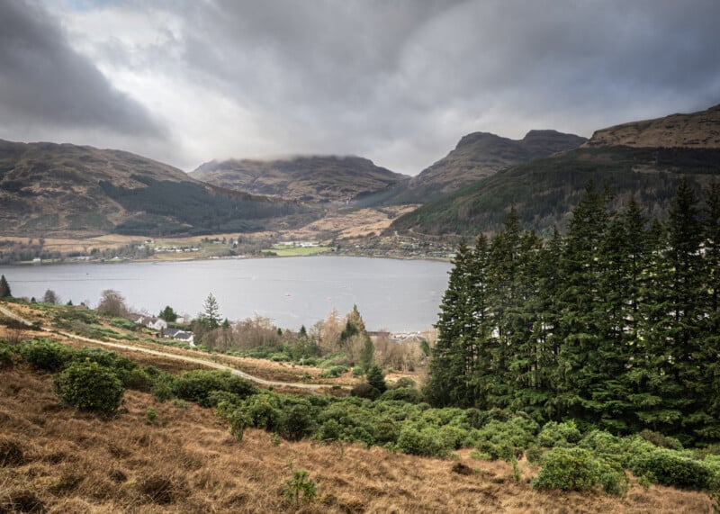 A cloudy sky hangs over a serene lake surrounded by mountains, dense evergreen trees, and patches of green and brown vegetation in a rural landscape.