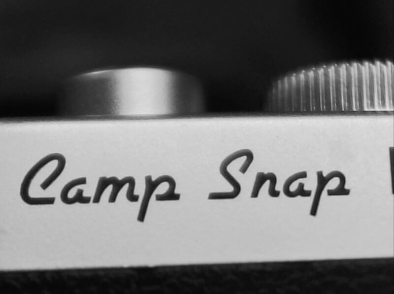 Close-up black and white photo of a camera with the words "Camp Snap" written in cursive on the body, alongside a textured dial and a smooth button.