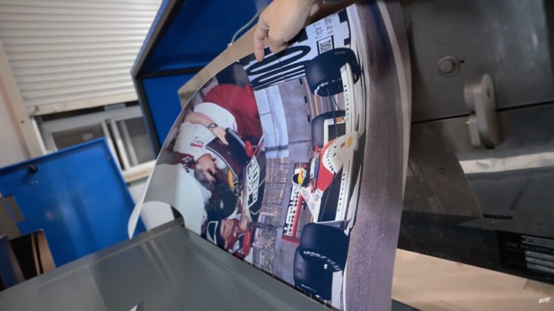 A hand pulls out a printed poster showing a racing car and two people next to it. The image comes out of a large blue and gray printer and is upside down from the viewer's perspective.