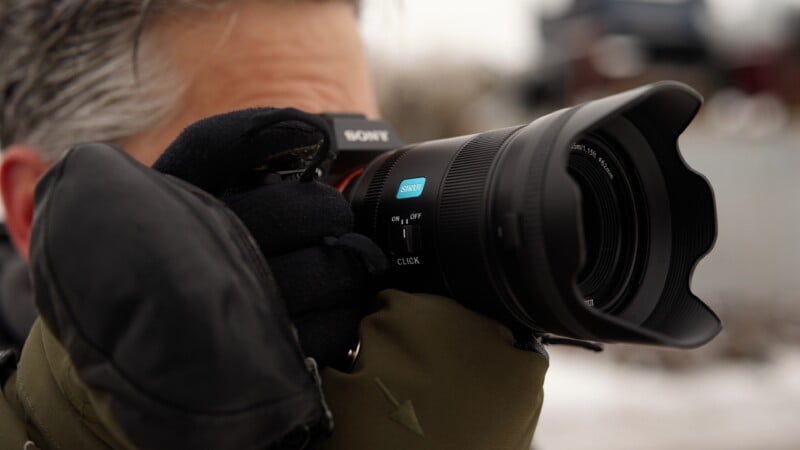Un hombre con guantes y una chaqueta verde sostiene una cámara Sony de lente grande cerca de su cara, preparándose para tomar una fotografía al aire libre en un día nublado.
