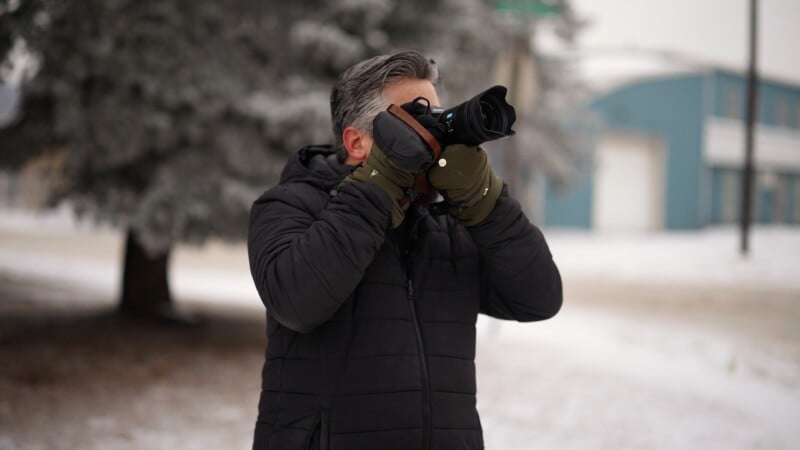 Un hombre vestido con un abrigo oscuro y guantes verdes sostiene una cámara frente a su cara y toma una fotografía al aire libre en la nieve con árboles y edificios azules al fondo.