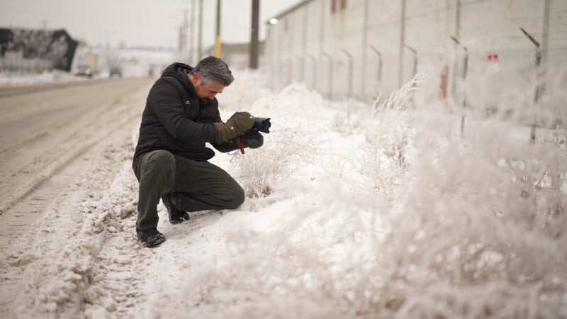 Un hombre vestido con una chaqueta negra y guantes se arrodilla al borde de una carretera nevada, fotografiando plantas cubiertas de escarcha con una cámara en un día nublado de invierno.