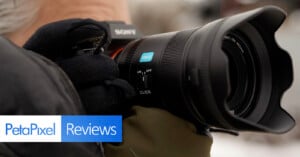 A close-up of a gloved person holding a Sony camera with a large lens. The image includes the "PetaPixel Reviews" logo in the bottom left corner.